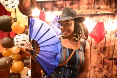 A woman with a black cowboy hat and cream beads in her hair poses with a paper folding fan in front of a wooden backdrop